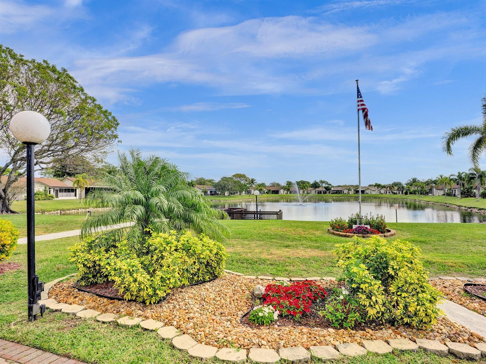Landscaped grounds featuring a stone-lined garden bed, mature greenery, a concrete walkway, and a pond with a fountain