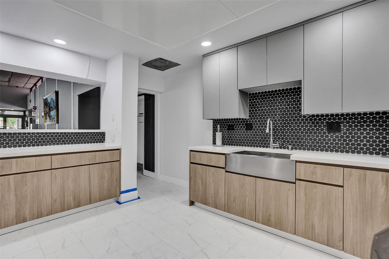 Flat-panel cabinetry in light wood-finish and matte gray, white stone-finish countertops, black penny-tile backsplash, stainless steel farmhouse sink, and large-format white tile flooring