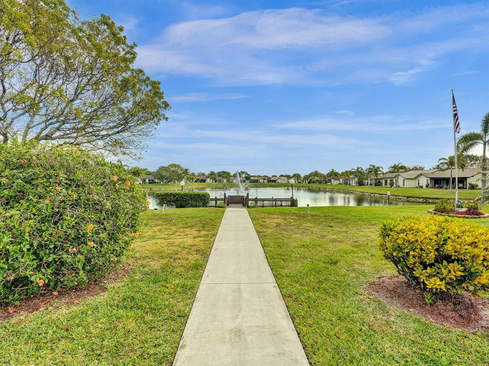 Concrete pathway leading to a pond with a fountain and wooden dock, surrounded by mature landscaping and lawn