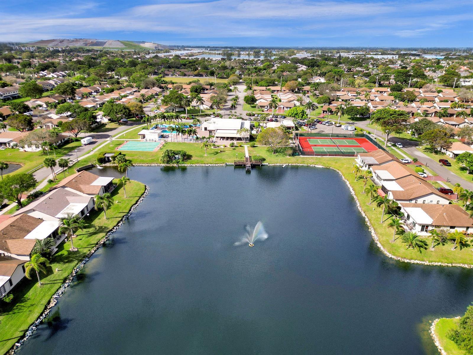 Aerial perspective of a community featuring a central lake with a fountain, tennis courts, swimming pool, and dedicated clubhouse facilities