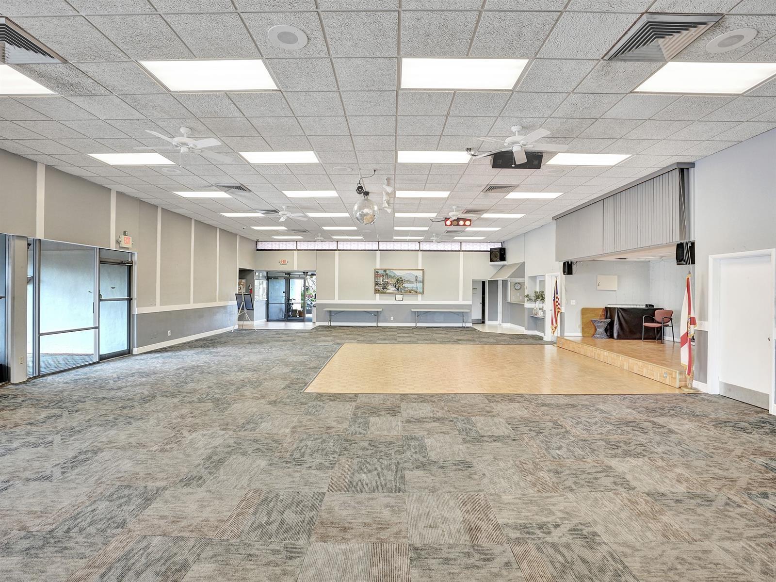 Open-concept interior featuring a raised wooden stage, integrated ceiling lighting, multiple overhead fans, and durable patterned carpeting