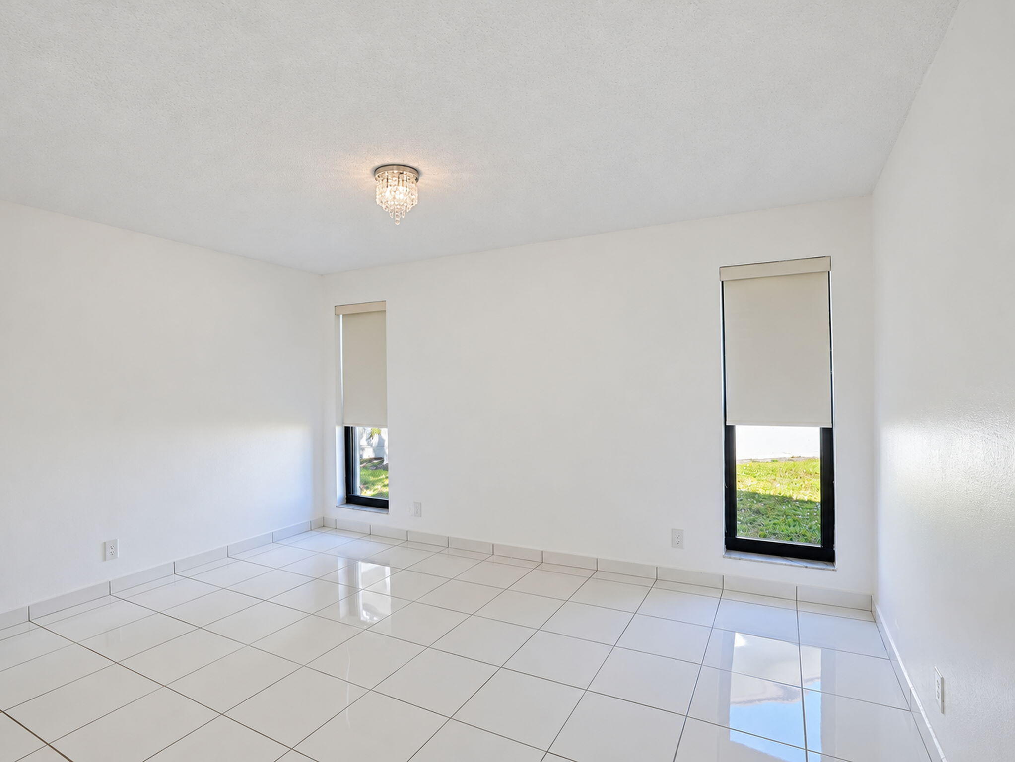 Neutral interior featuring large-format white tile flooring, dual floor-to-ceiling windows, roller shades, and a flush-mount crystal light fixture