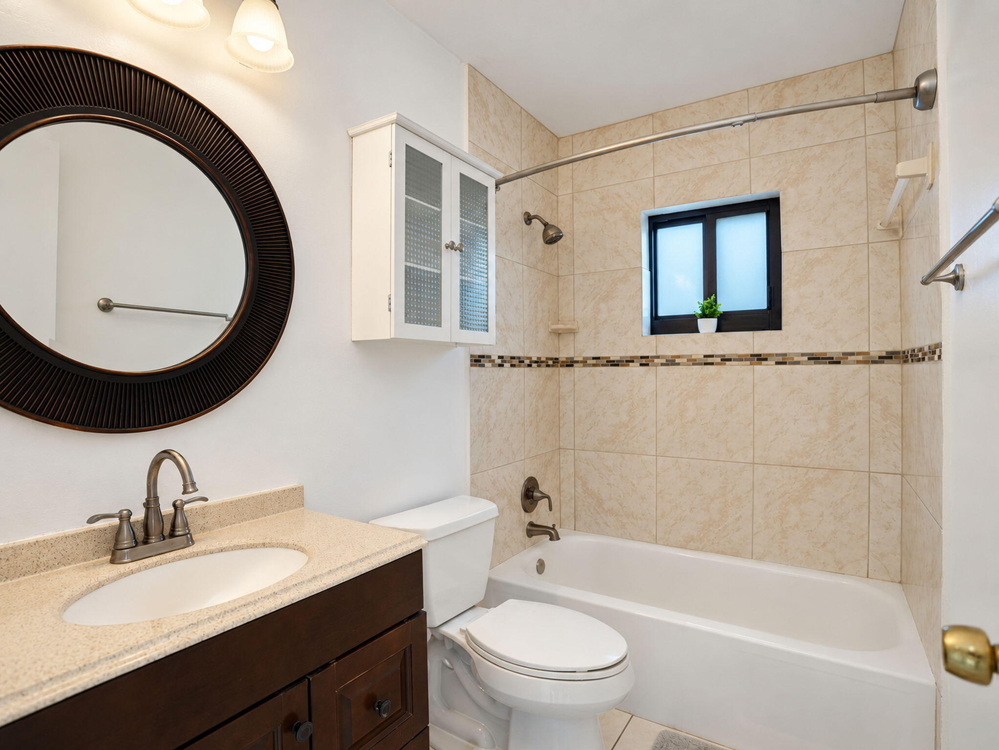 Bathroom featuring a dark wood vanity with a solid surface countertop, oval undermount sink, ceramic tile shower surround with decorative mosaic accent, and a white wall-mounted cabinet