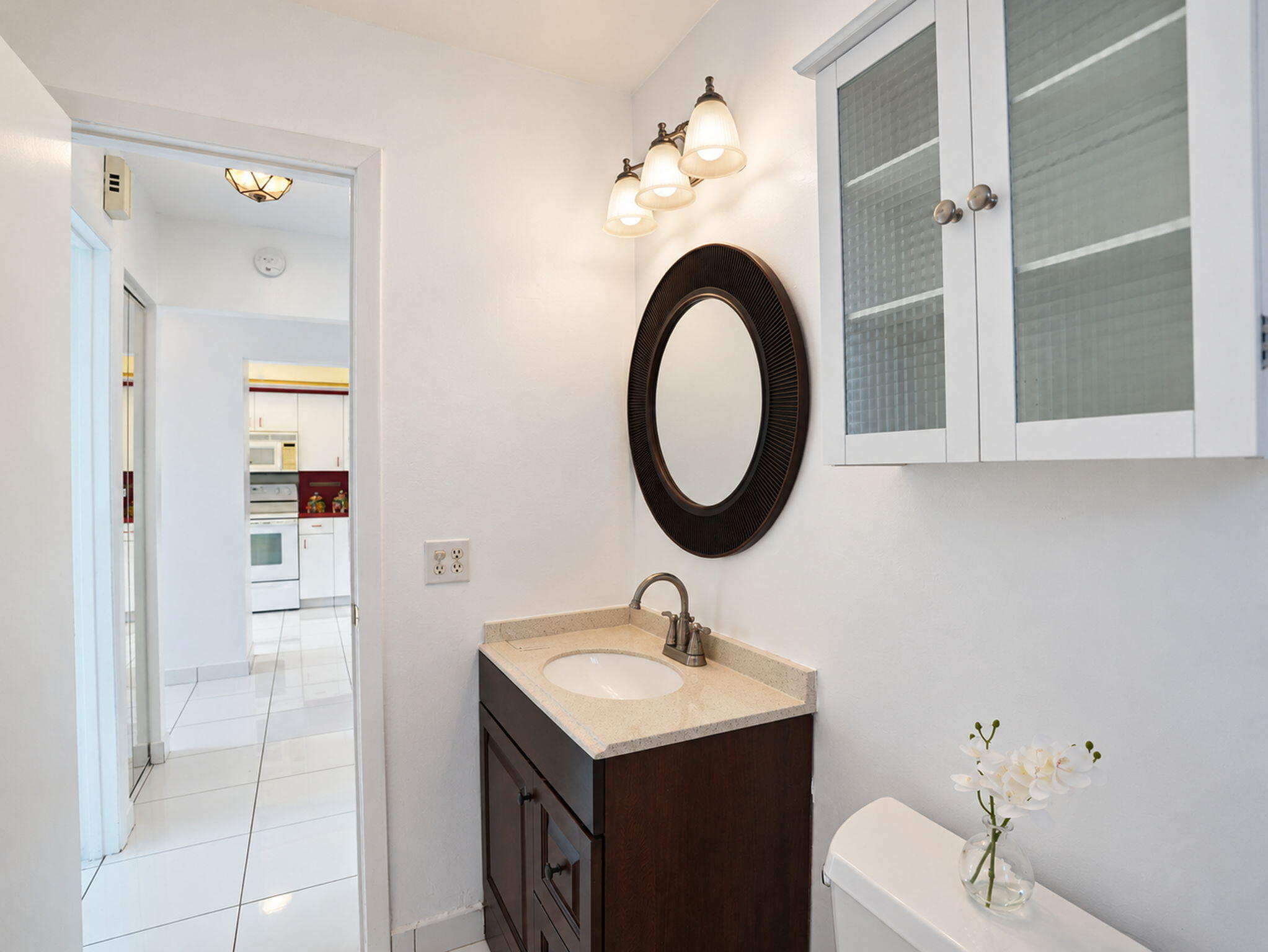 Dark wood vanity featuring an oval mirror, neutral stone countertop with an integrated basin, three-light fixture, and elevated wall-mounted cabinet with textured glass panels