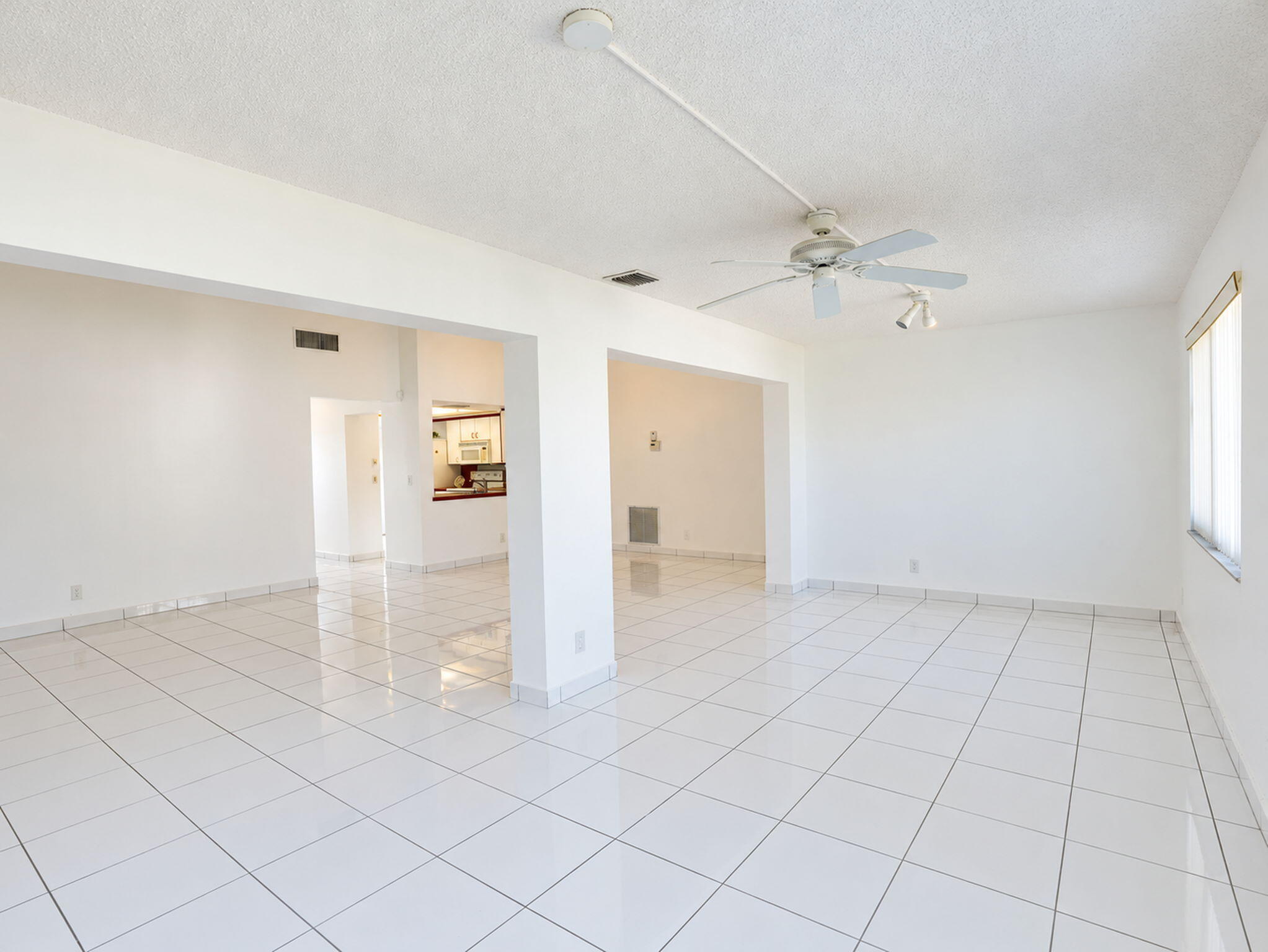 Open-concept interior featuring large-format white tile flooring, structural support pillars, a ceiling fan with integrated lighting, and horizontal window blinds