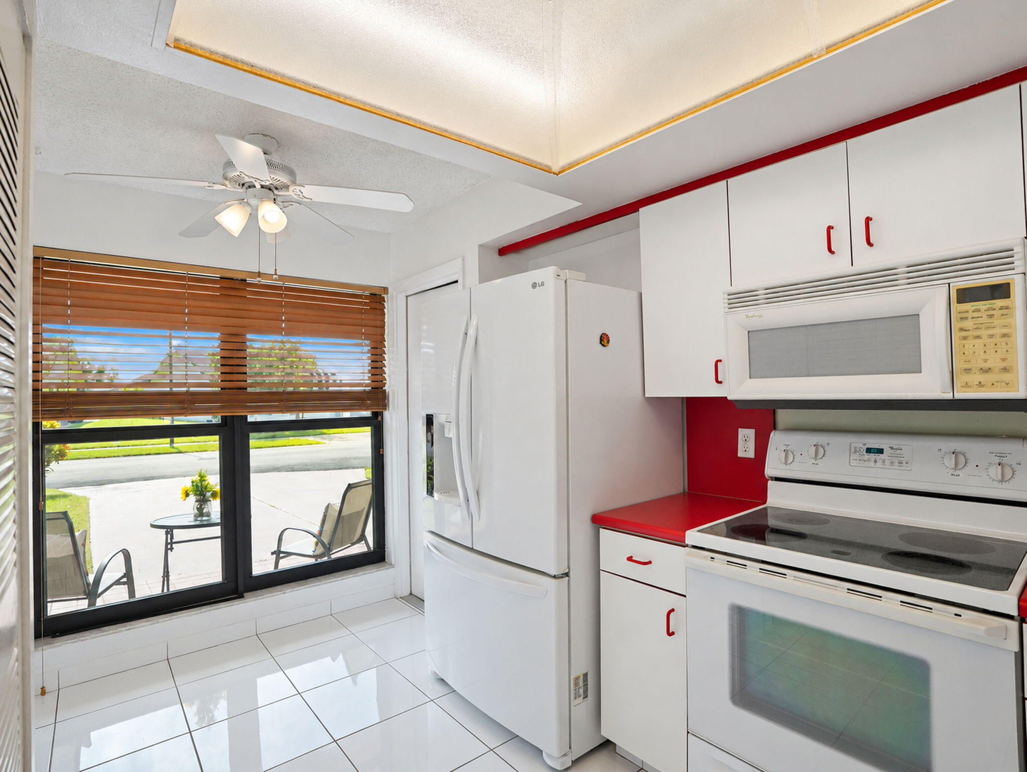Kitchen featuring white cabinetry, contrasting red countertops, tiled flooring, a tray ceiling with integrated lighting, and a large window with wood-finish blinds