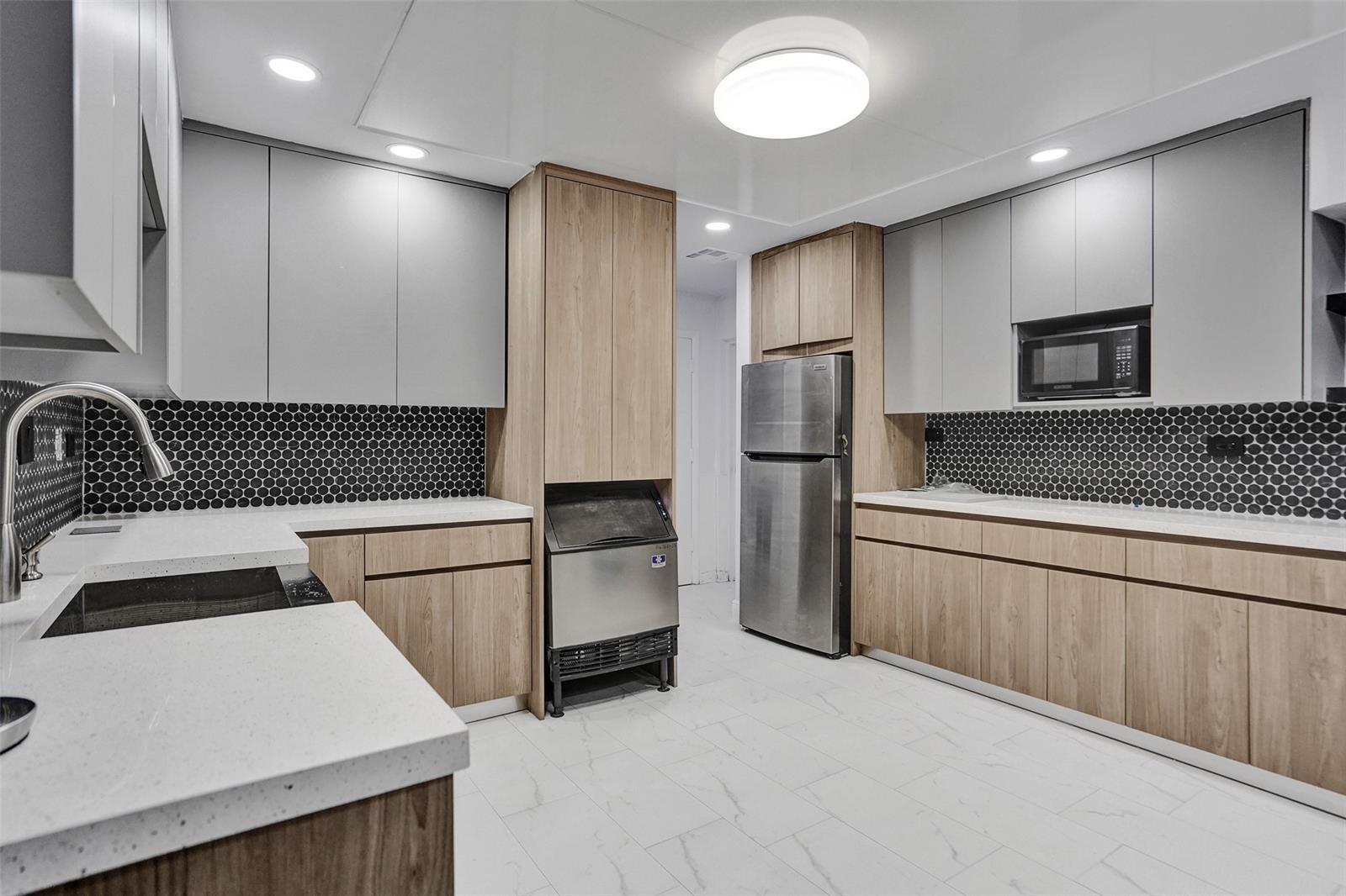 Kitchen featuring two-tone cabinetry with wood-finish lower and light-toned upper units, solid-surface countertops, black hexagonal tile backsplash, stainless steel appliances, and large-format tile flooring