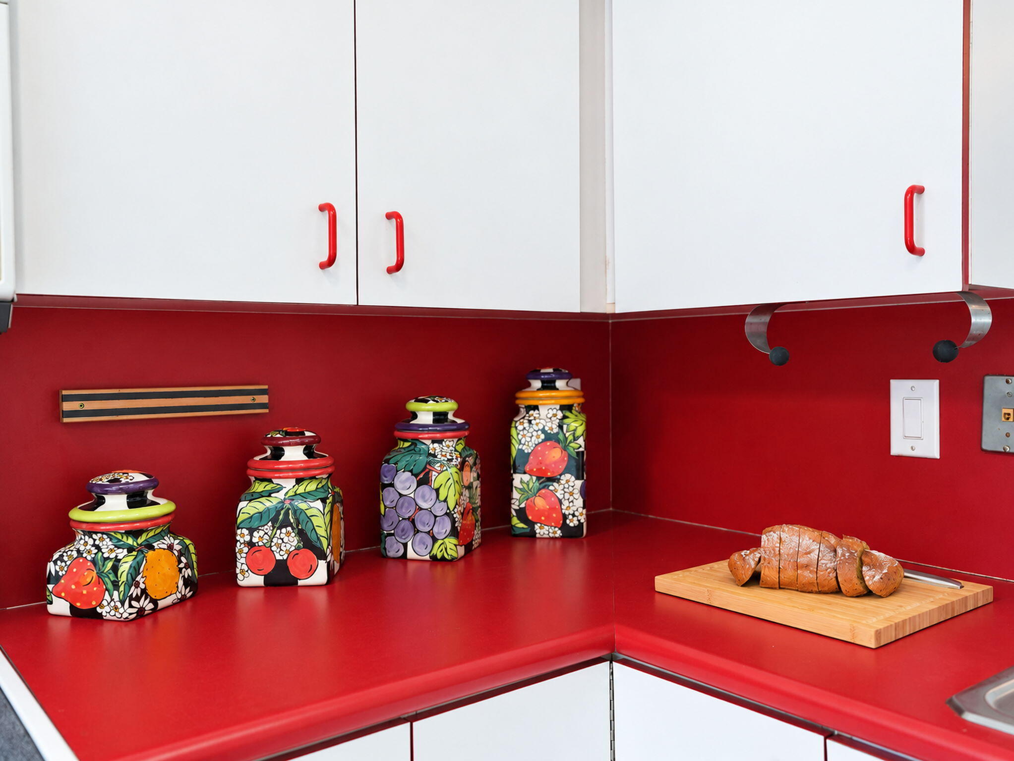 White cabinetry featuring red hardware, complemented by a solid-surface red countertop and matching backsplash with an integrated wall-mounted magnetic knife rack