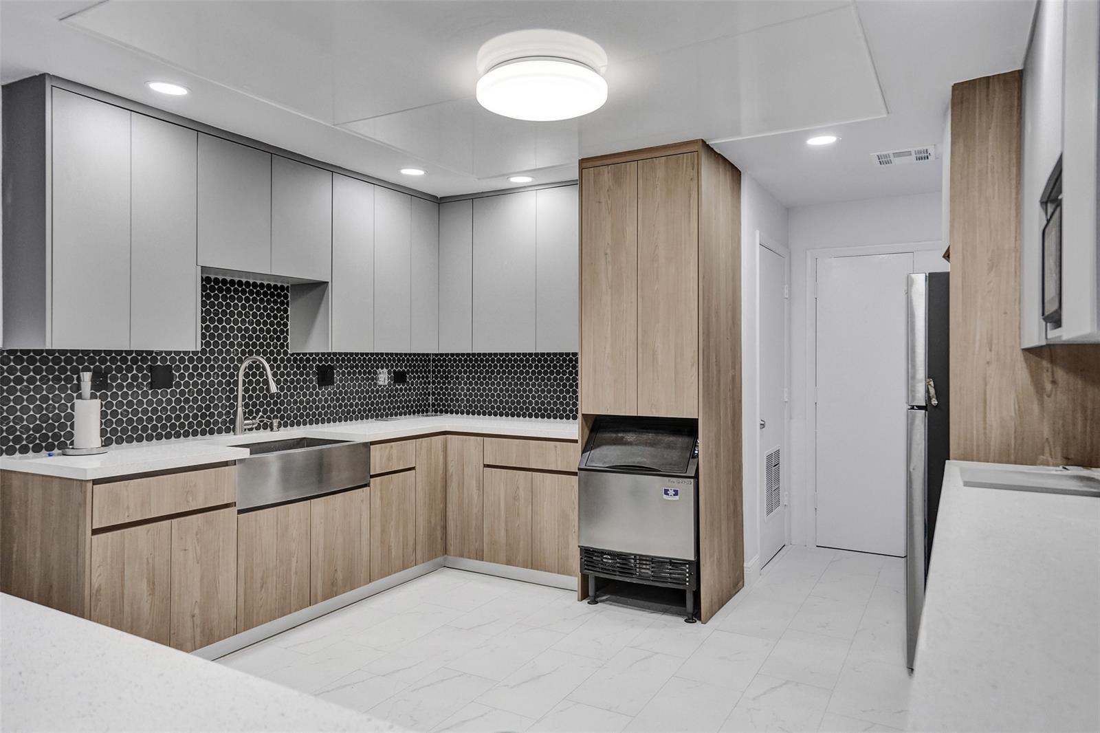 Kitchen featuring two-tone cabinetry with wood-finish lower units and light gray upper units, stone-surface countertops, stainless steel farmhouse sink, and a black hexagonal tile backsplash