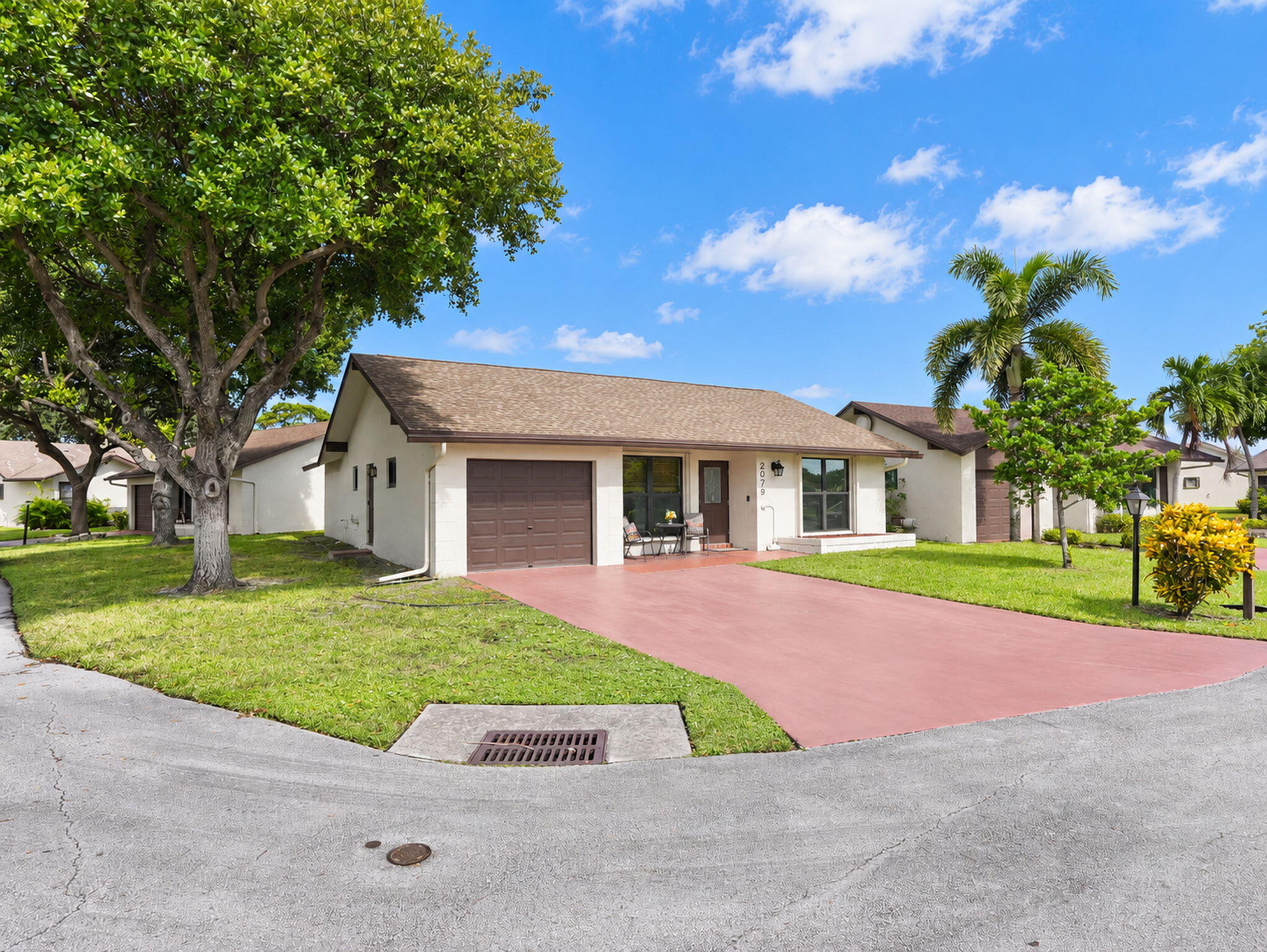 Single-story exterior featuring a shingled roof, attached one-car garage, red-toned paved driveway, and mature landscaping