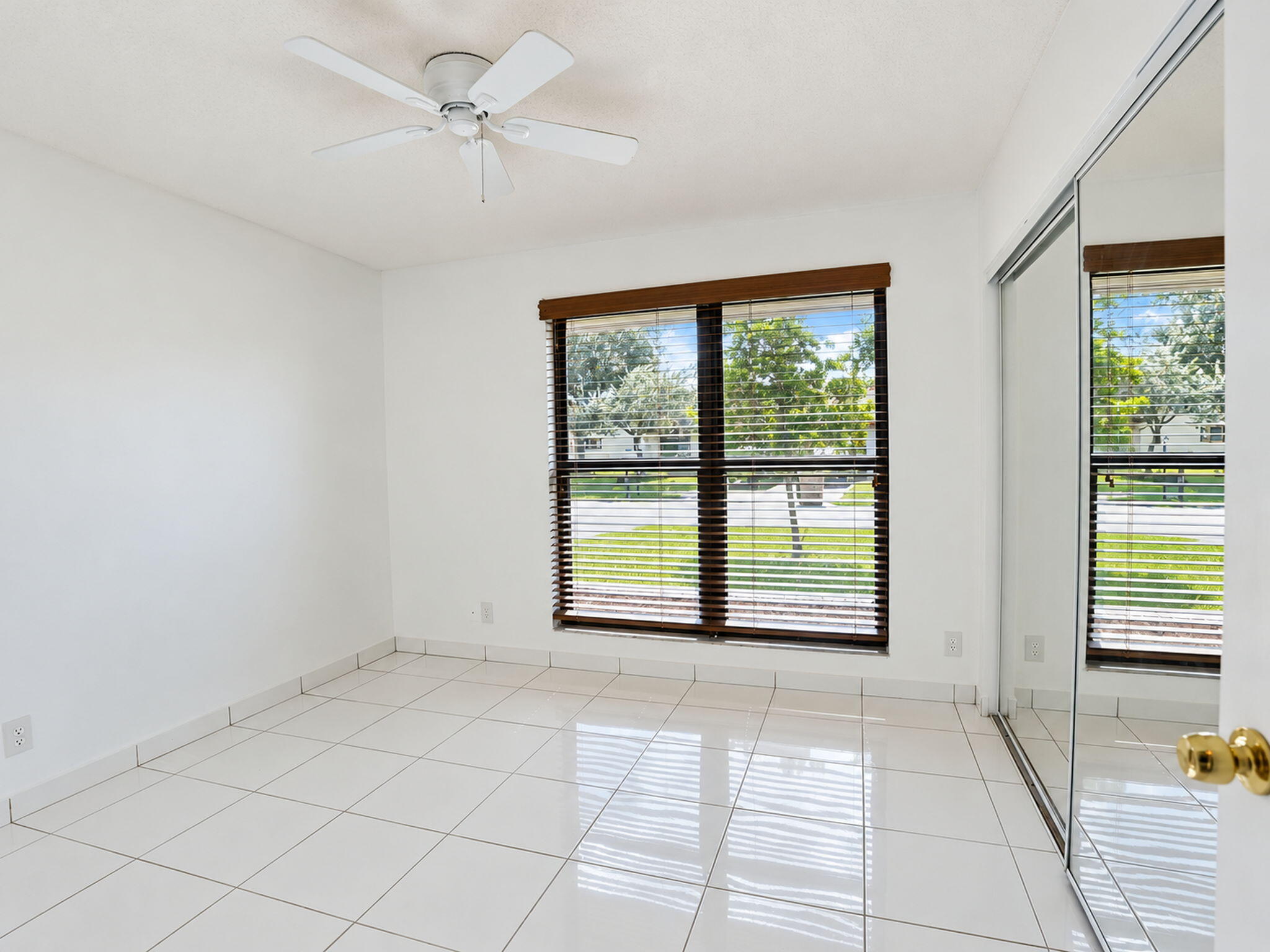 Neutral tile flooring, ceiling fan, double-hung window with dark wood-finish blinds, and floor-to-ceiling mirrored closet doors