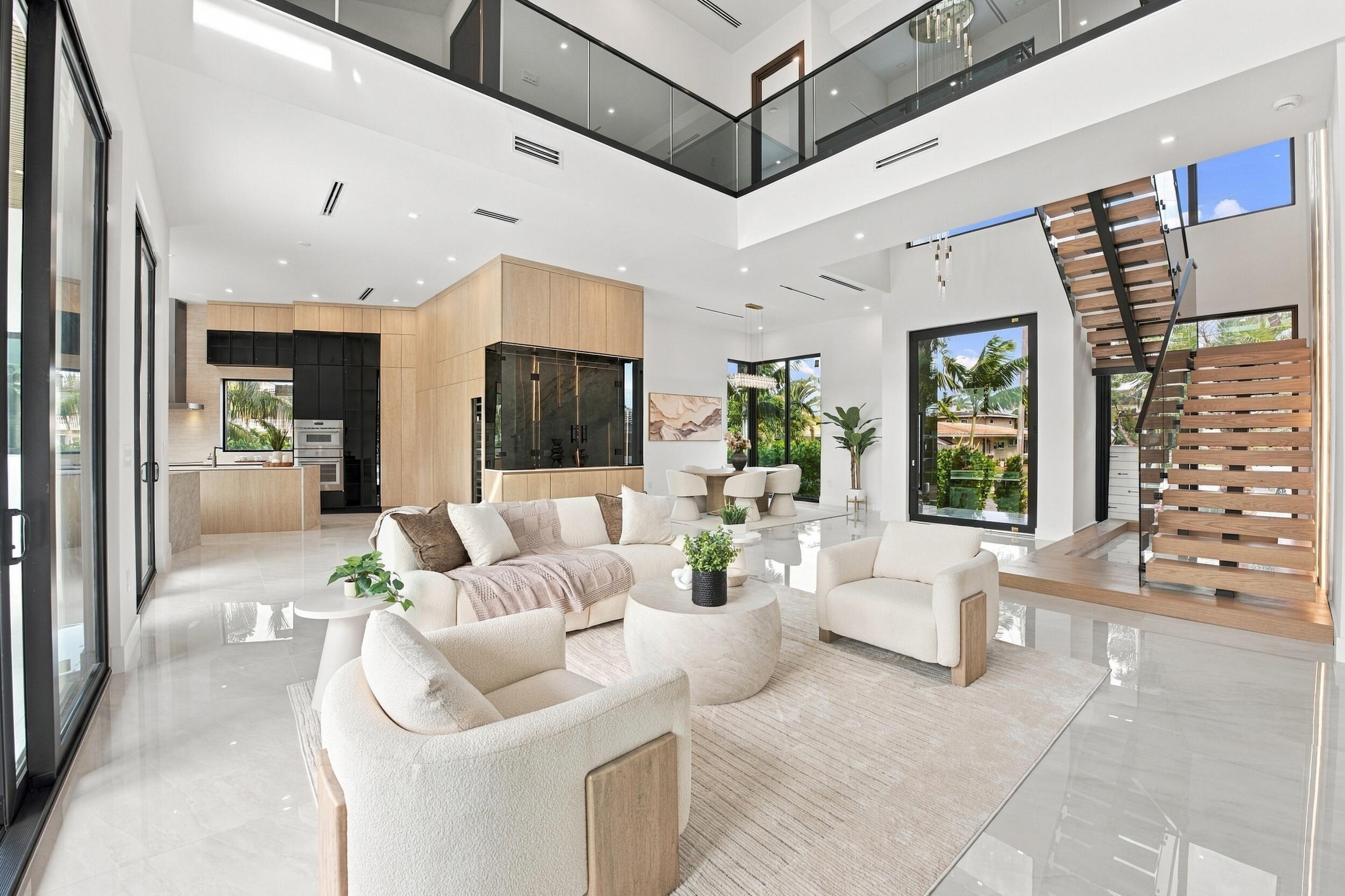 Open-concept interior featuring double-height ceilings, a wood-tread staircase with glass railing, large-format polished tile flooring, floor-to-ceiling cabinetry, and expansive glass doors