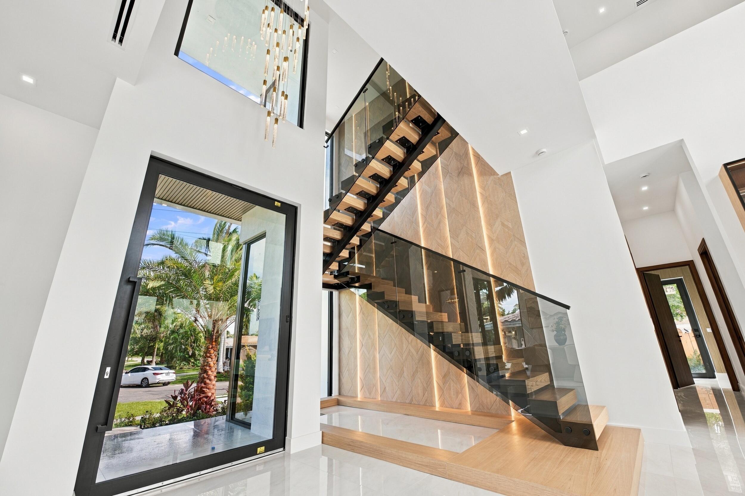 Double-height entryway featuring a floating wood staircase with glass balustrades, large-format polished flooring, a floor-to-ceiling accent wall with integrated vertical lighting, and a prominent glass pivot entry door