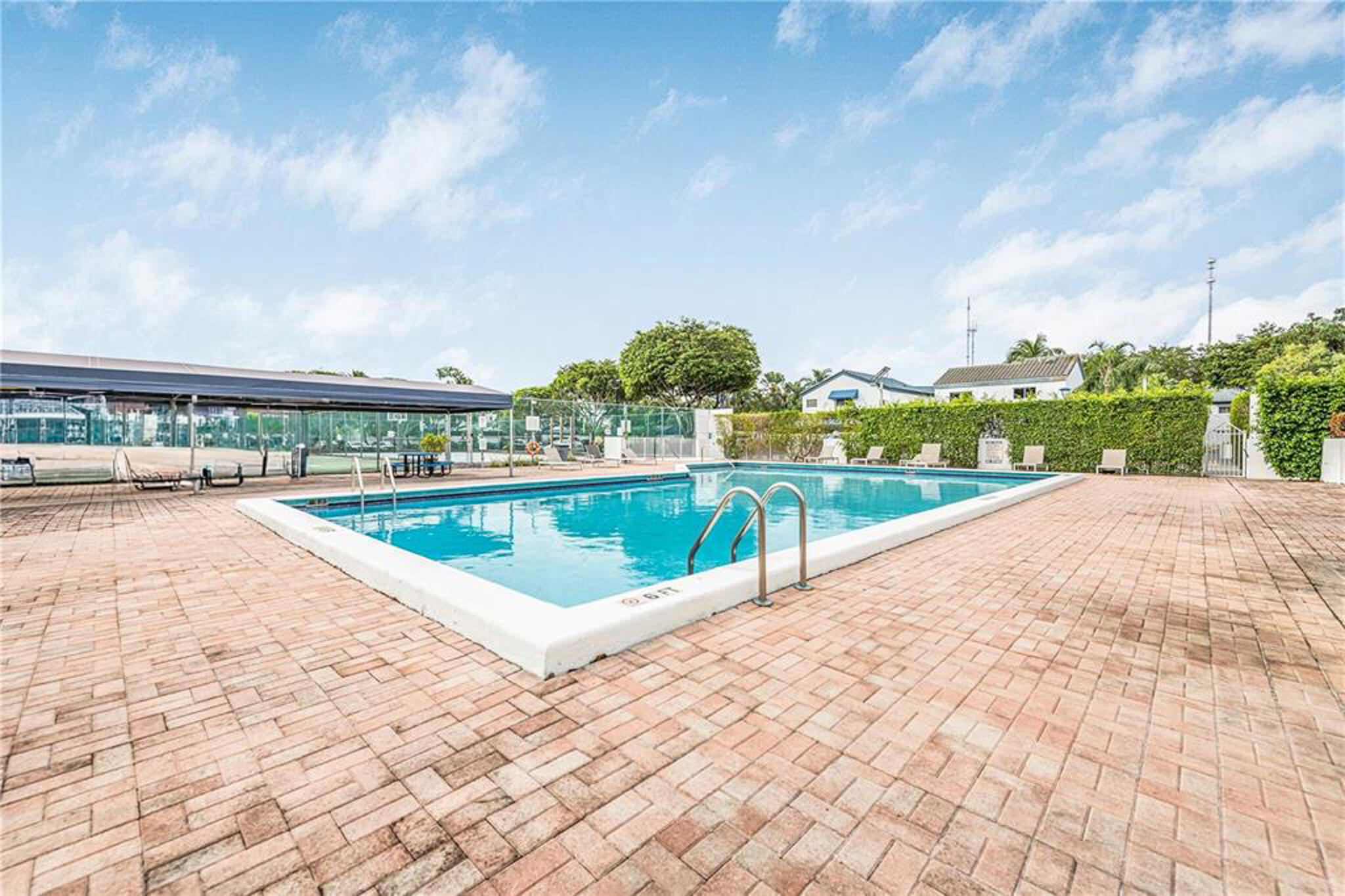 Outdoor swimming pool featuring a brick-paved deck, perimeter privacy hedging, and an attached covered structure