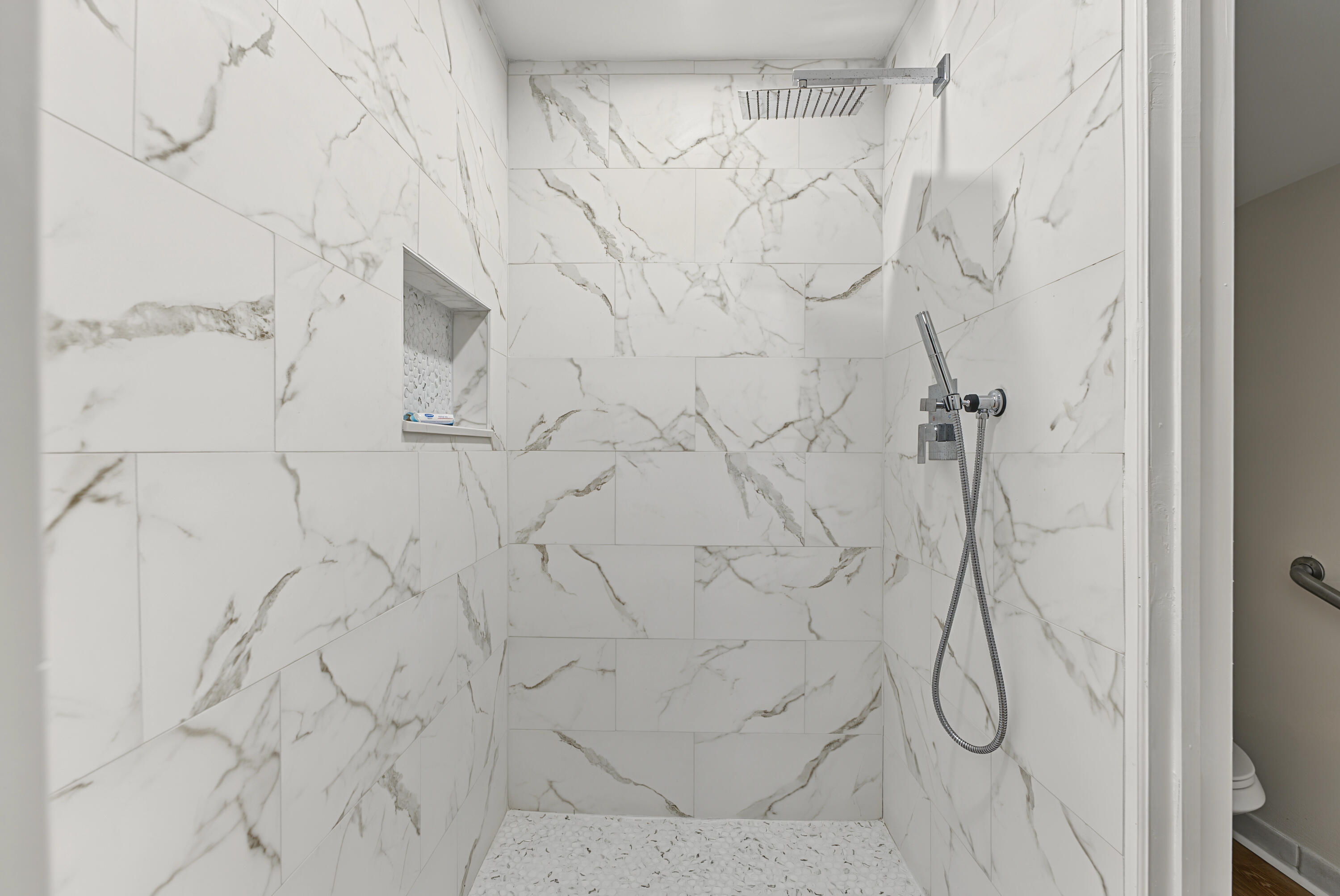 Walk-in shower featuring large-format white stone-finish wall tiling with grey veining, a built-in recessed niche, a ceiling-mounted rainfall showerhead, a separate handheld shower wand, and a textured white floor tiling