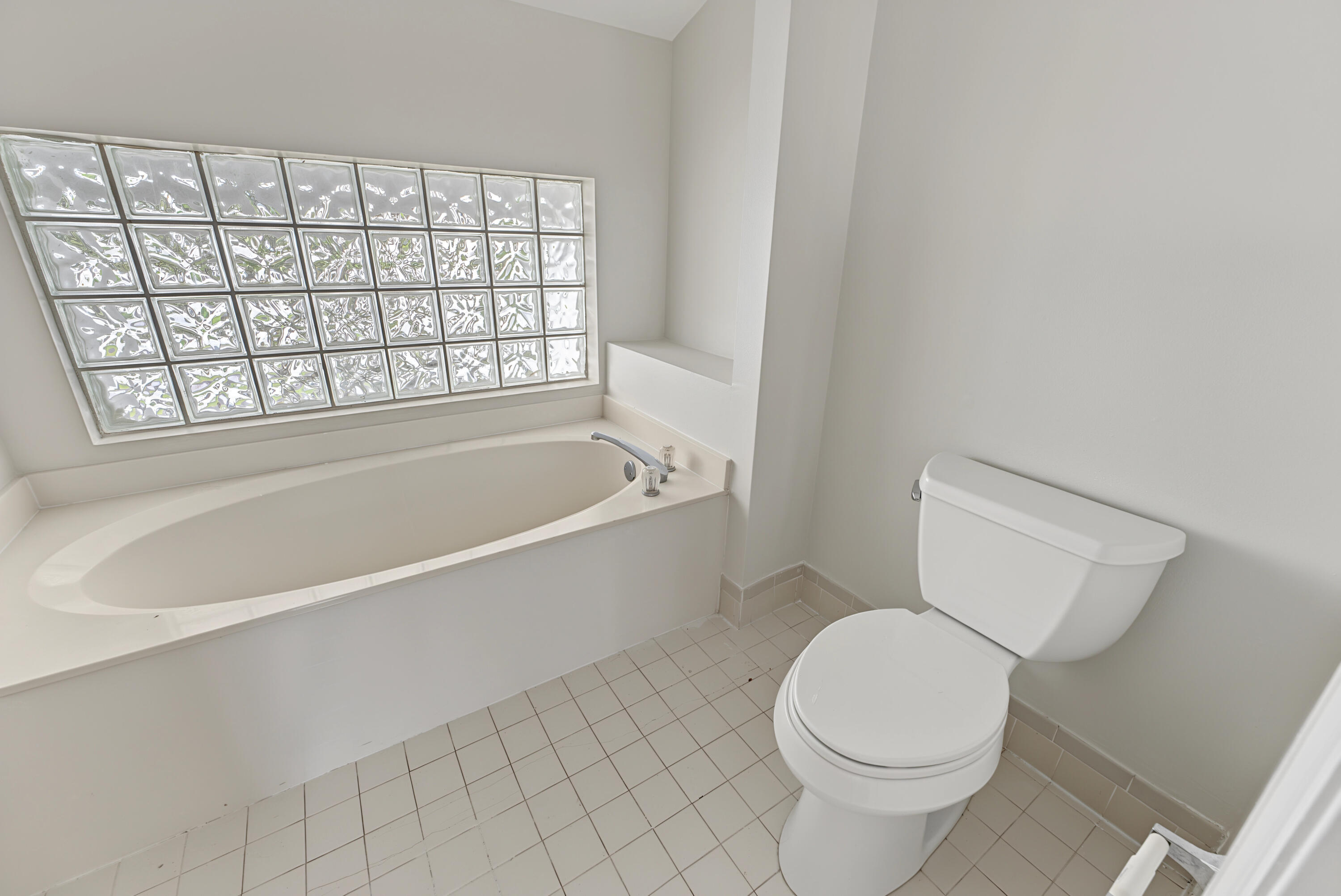 Oval soaking tub with integrated deck, glass block window, chrome plumbing fixtures, and ceramic tile flooring