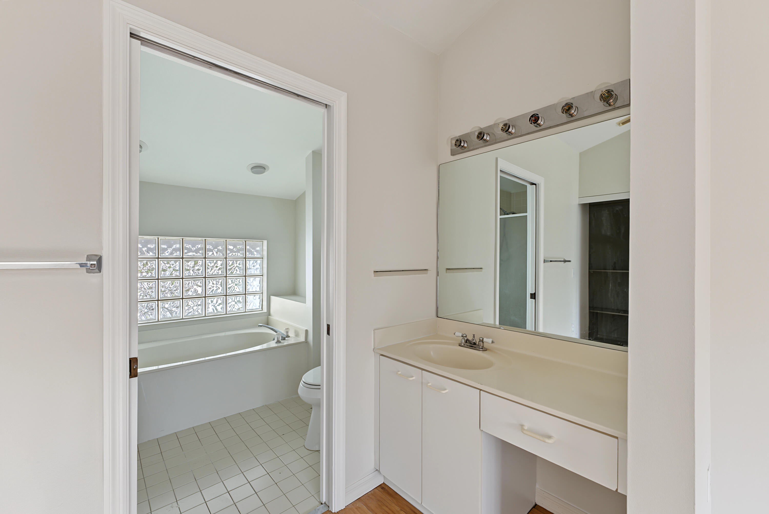 Bathroom featuring white cabinetry, integrated countertop with oval sink, large rectangular mirror, and multi-bulb vanity light, with a secondary space containing a built-in soaking tub and glass block window