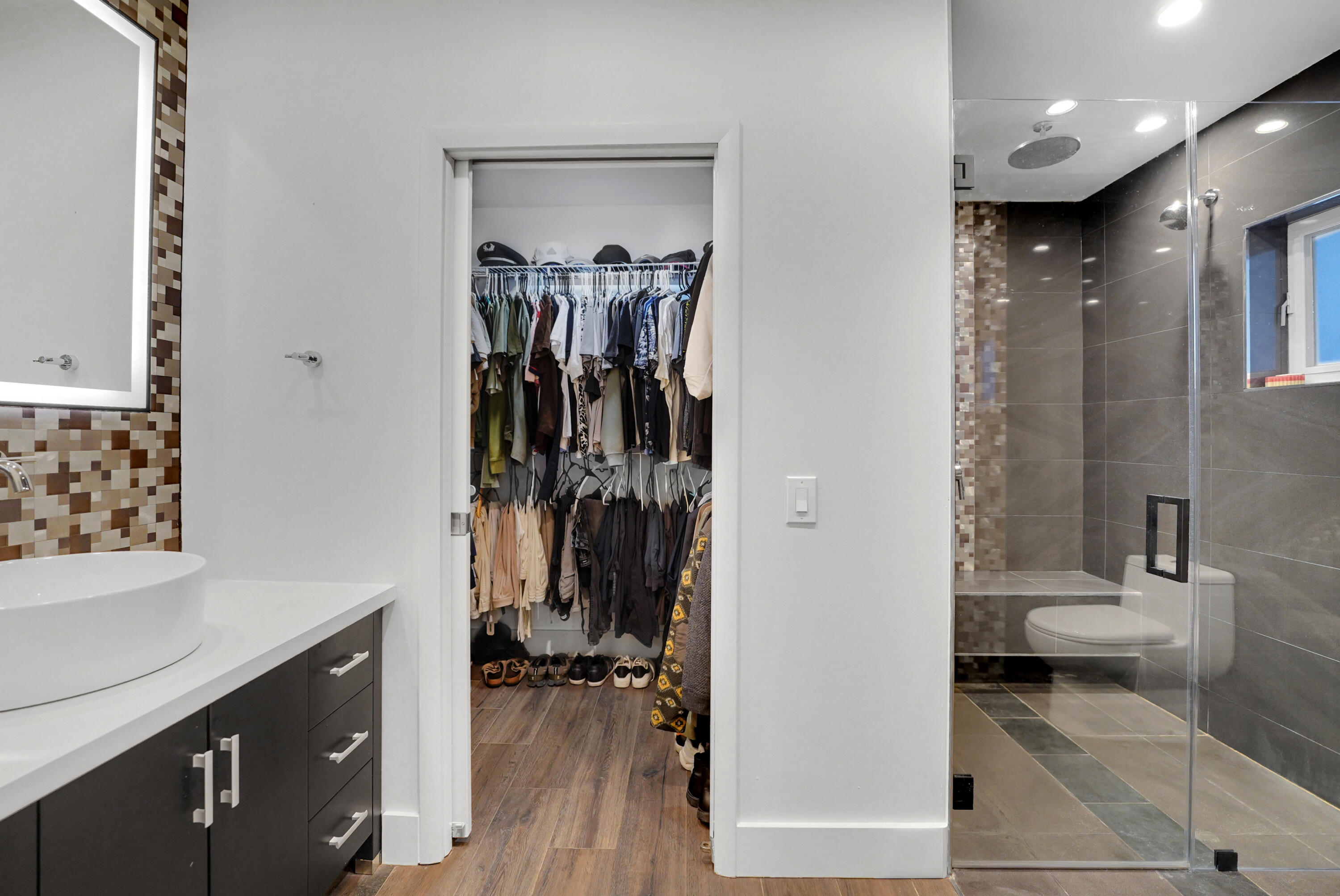 Bathroom featuring a dark wood-finish vanity with a white countertop and vessel sink, illuminated mirror, walk-in closet with built-in wire shelving, and a glass-enclosed shower with large-format tile and built-in bench