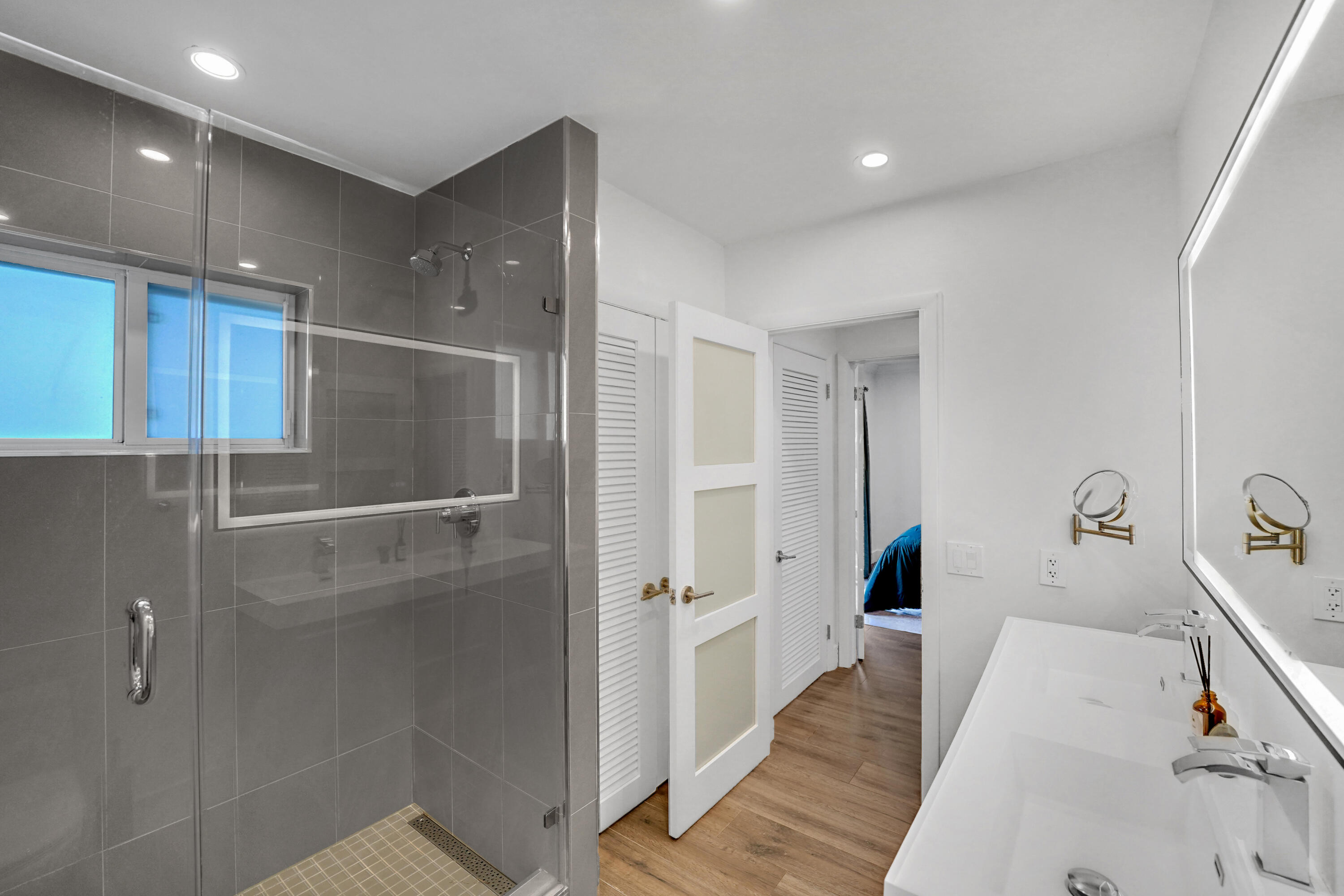 Walk-in shower with grey tile surround, dual-sink vanity, backlit vanity mirror, recessed lighting.