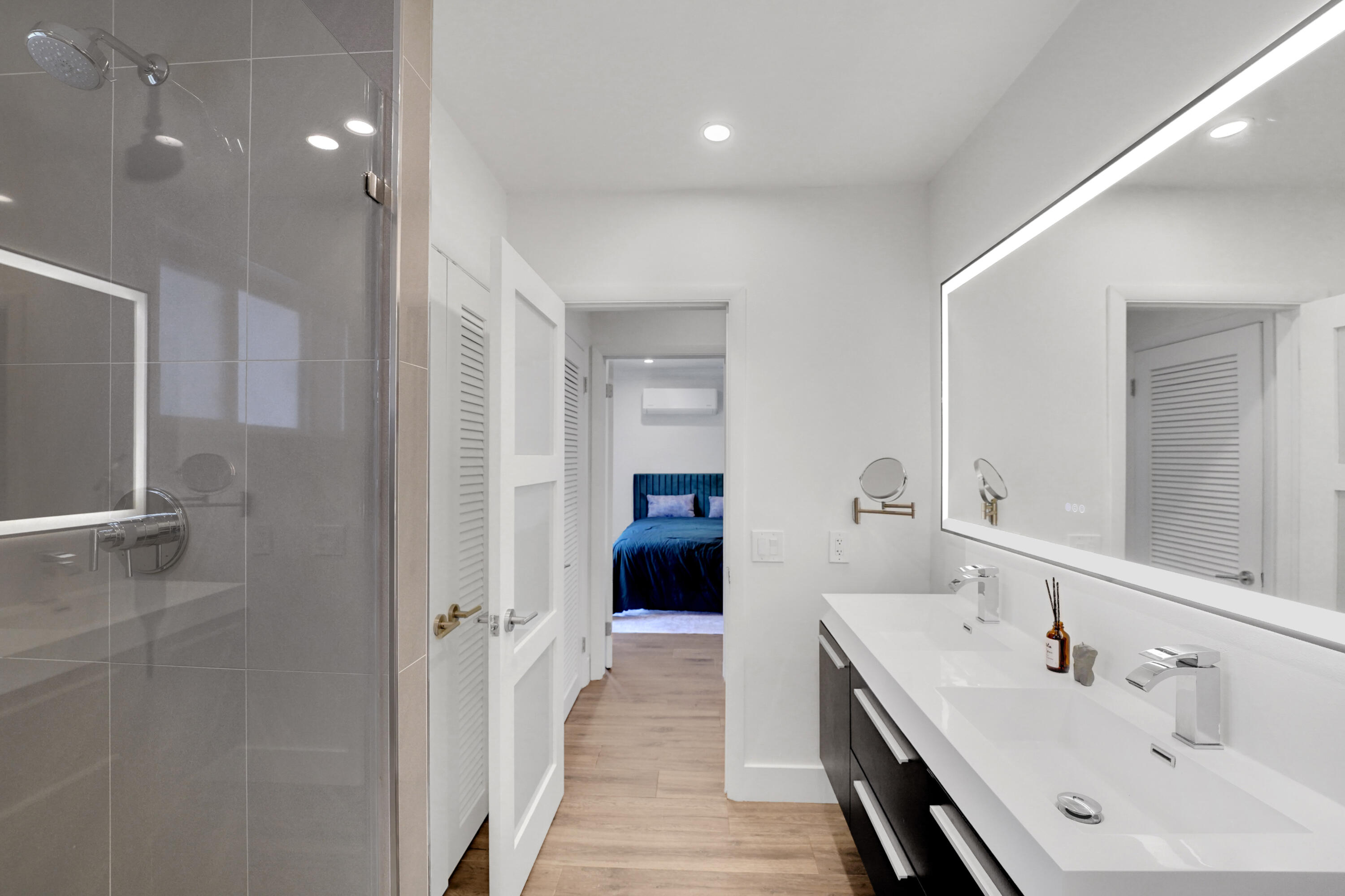 Dual-sink floating vanity with white countertops, expansive backlit mirror, glass-enclosed shower with large-format tile surround.