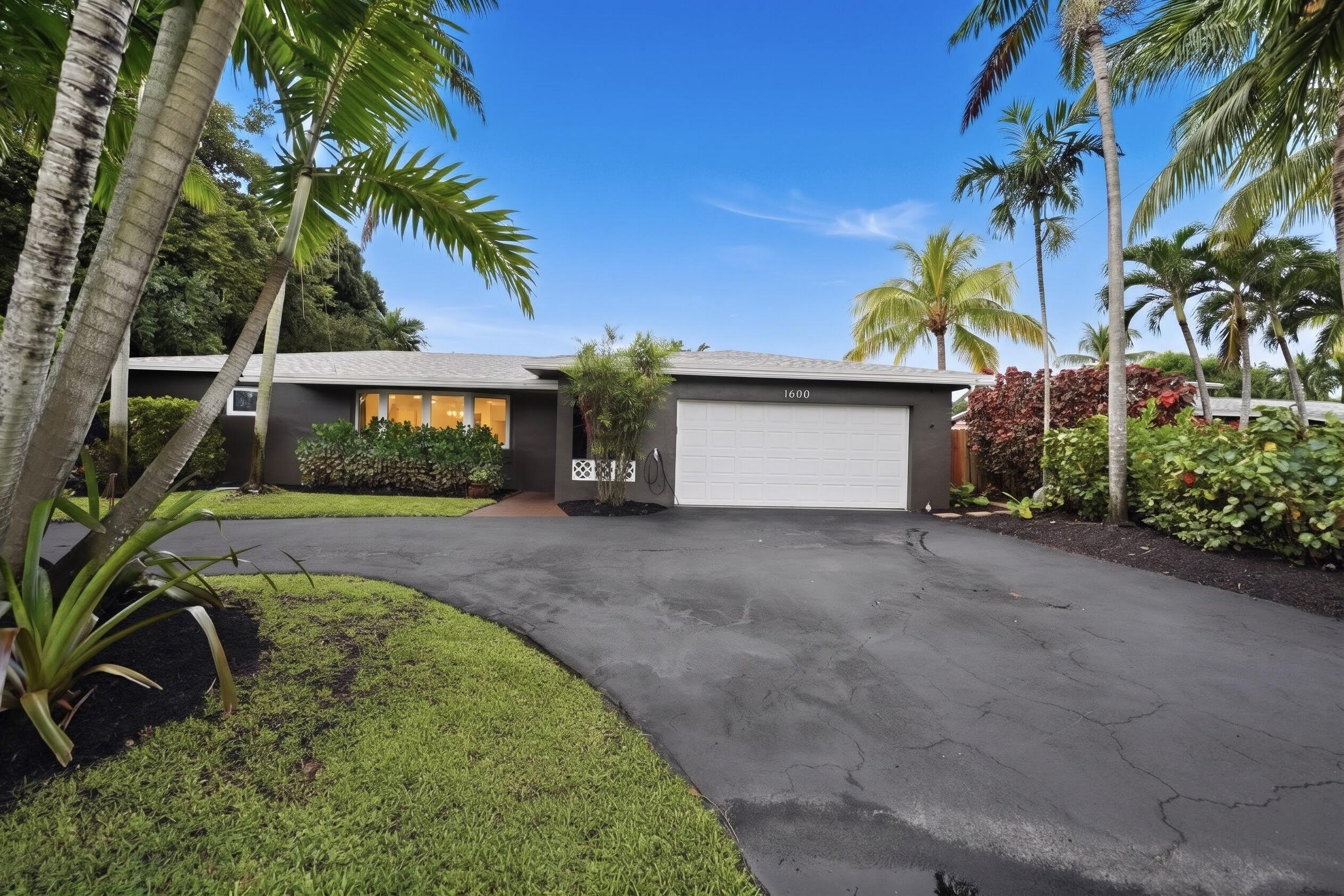 Single-story exterior featuring a dark-toned facade, an attached two-car garage, a curved asphalt driveway, and mature landscaping including palm trees