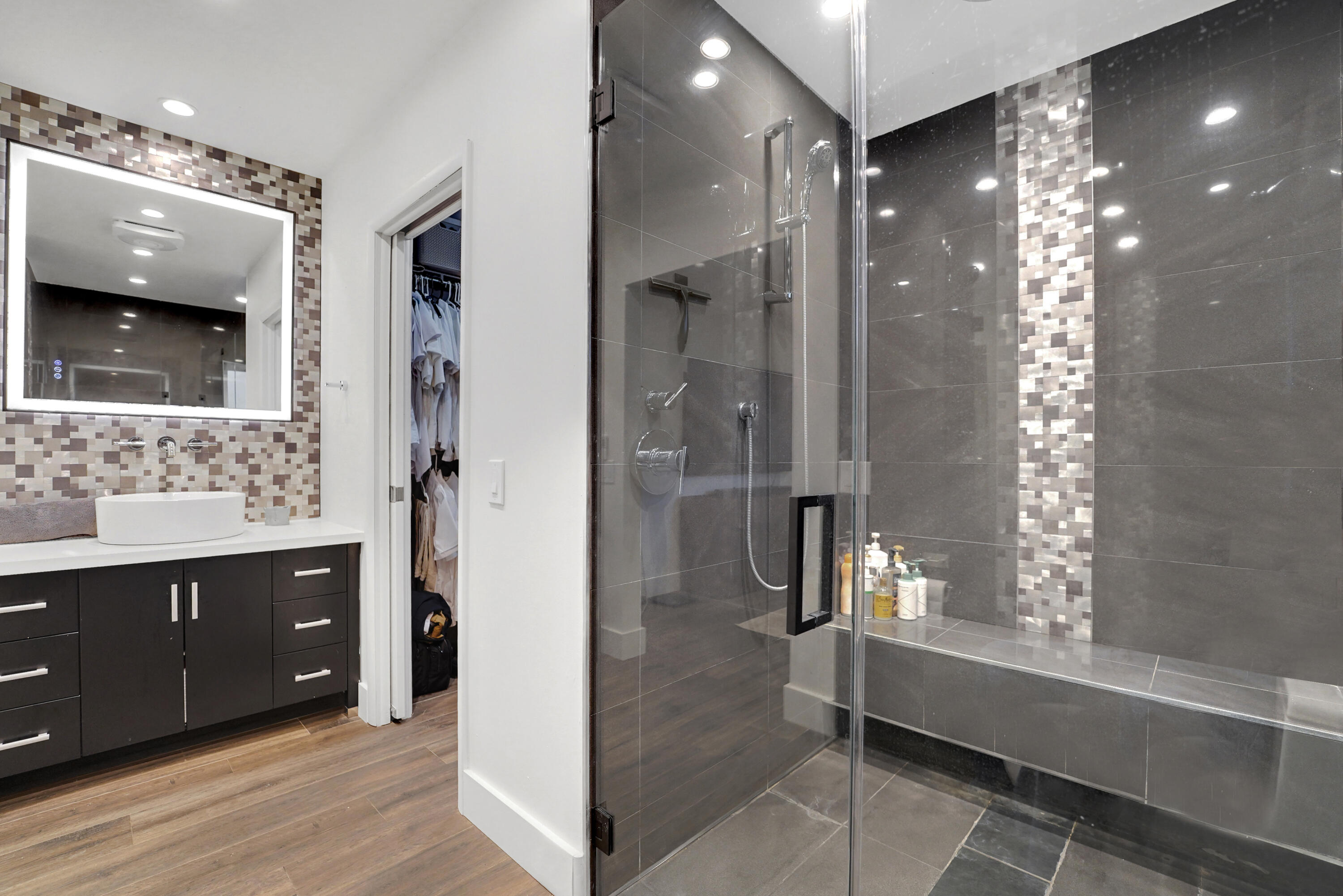 Bathroom featuring a dark wood-finish vanity with a vessel sink, a backlit mirror, wood-finish flooring, and a glass-enclosed walk-in shower with tile detailing and a built-in bench