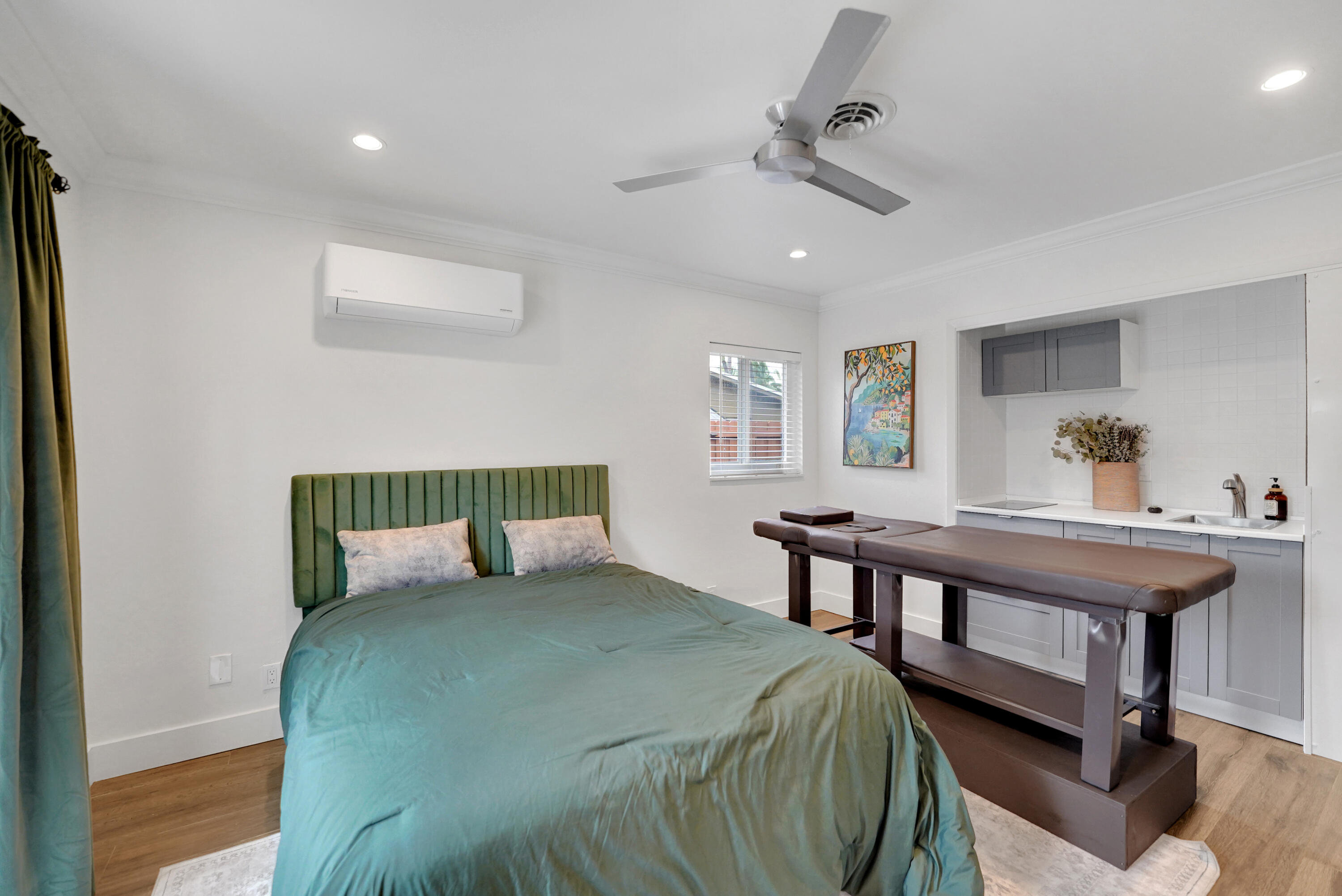 Room featuring wood-finish flooring, a ceiling fan, a wall-mounted climate control unit, and a built-in kitchenette with grey cabinetry and an integrated sink