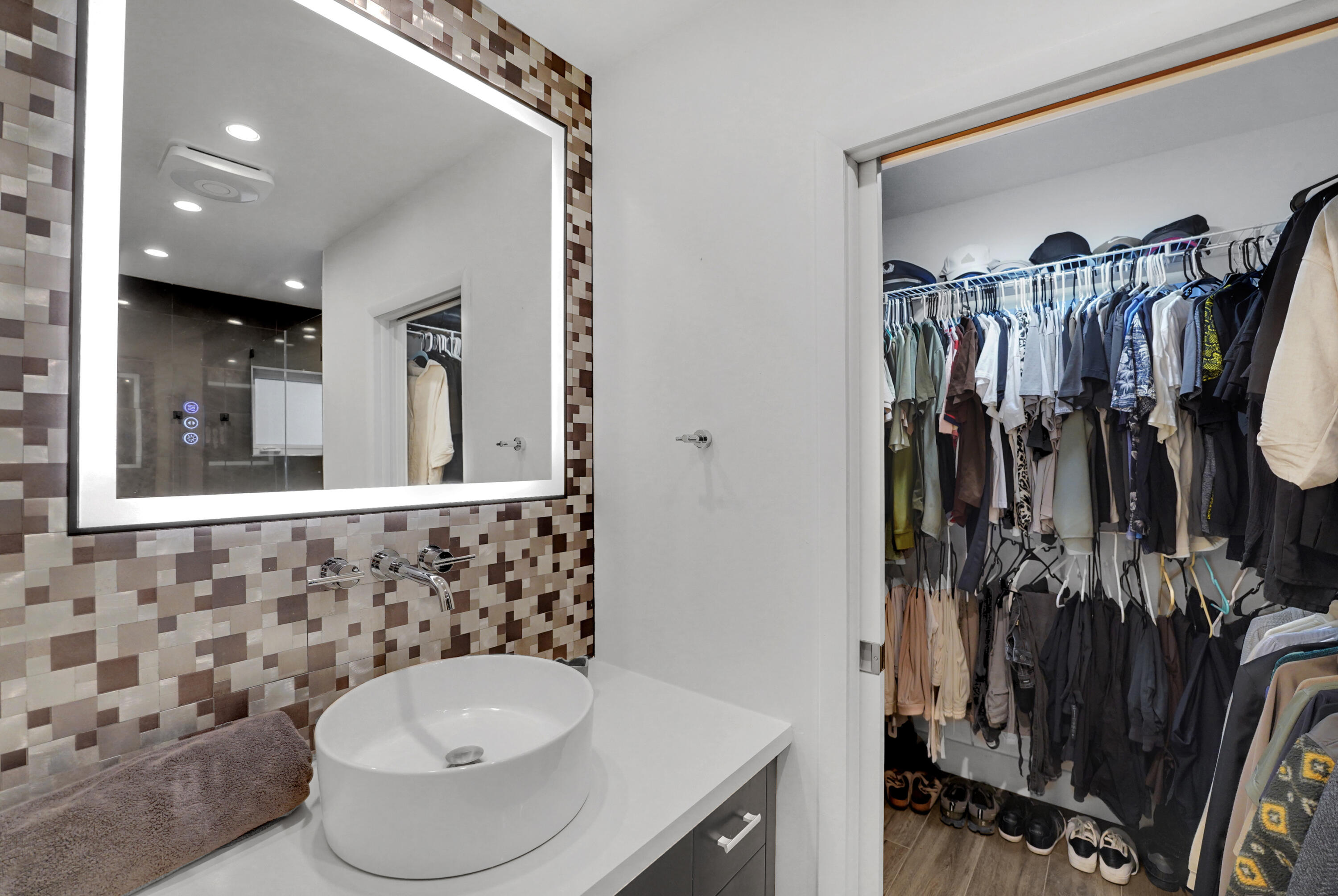 Modern bathroom featuring a rectangular vessel sink, wall-mounted faucet, illuminated mirror with integrated lighting, custom mosaic-tile backsplash, and wire-rack shelving system