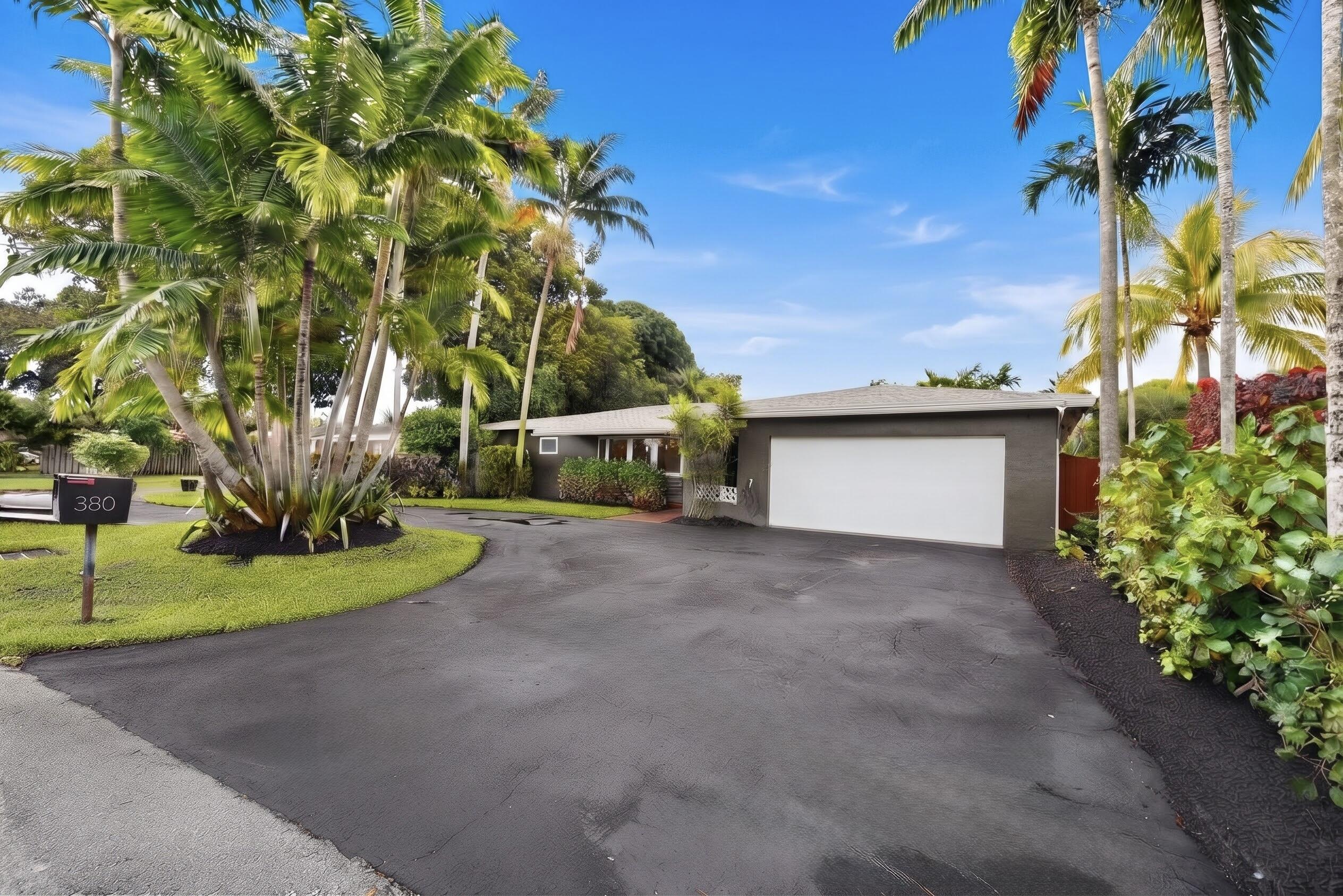 Single-story residence featuring a painted brick exterior, an attached two-car garage, an expansive asphalt driveway, and mature tropical landscaping