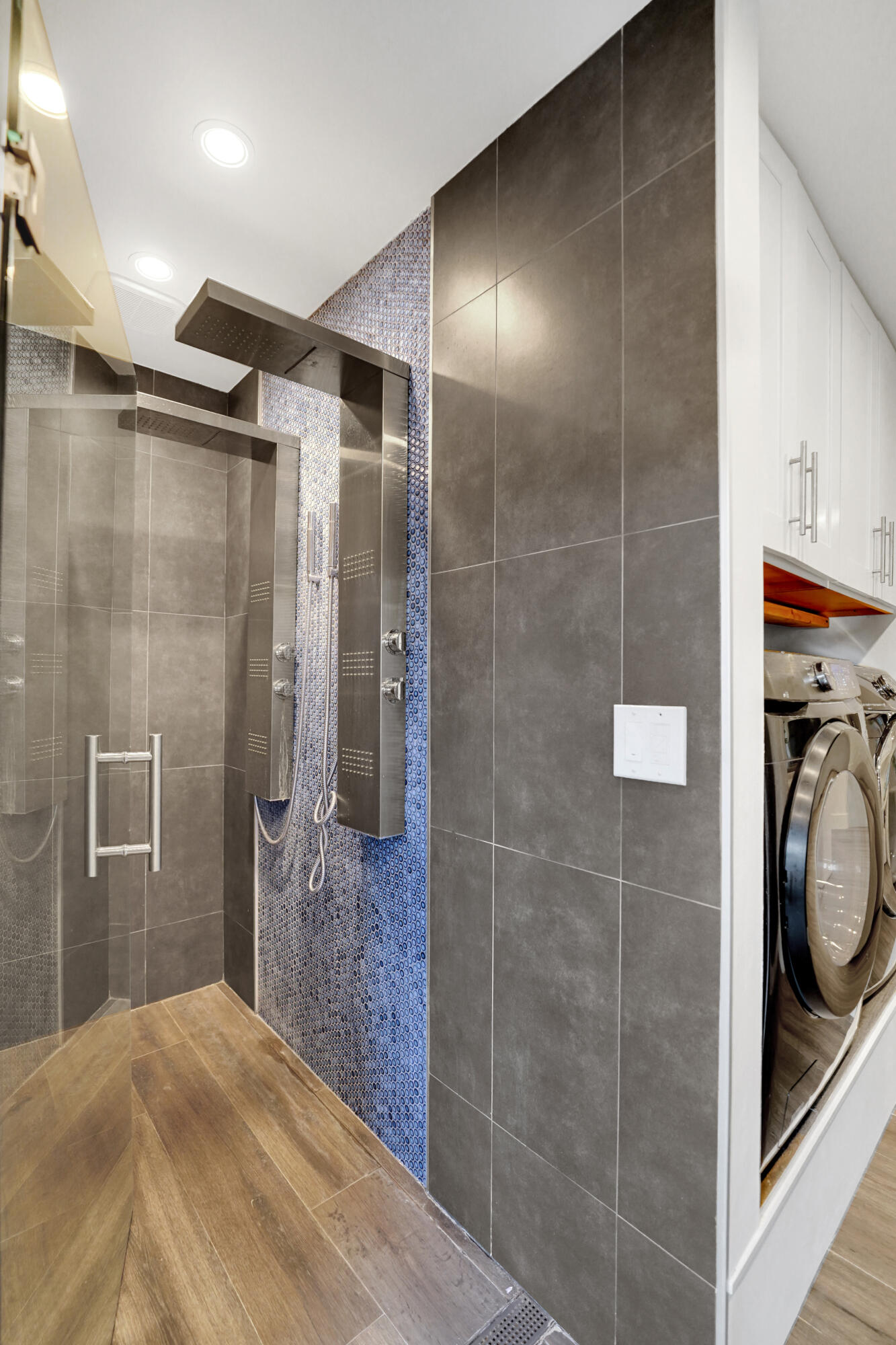 Walk-in shower featuring a metallic shower panel, blue mosaic tile accents, large-format grey wall tile, glass enclosure.