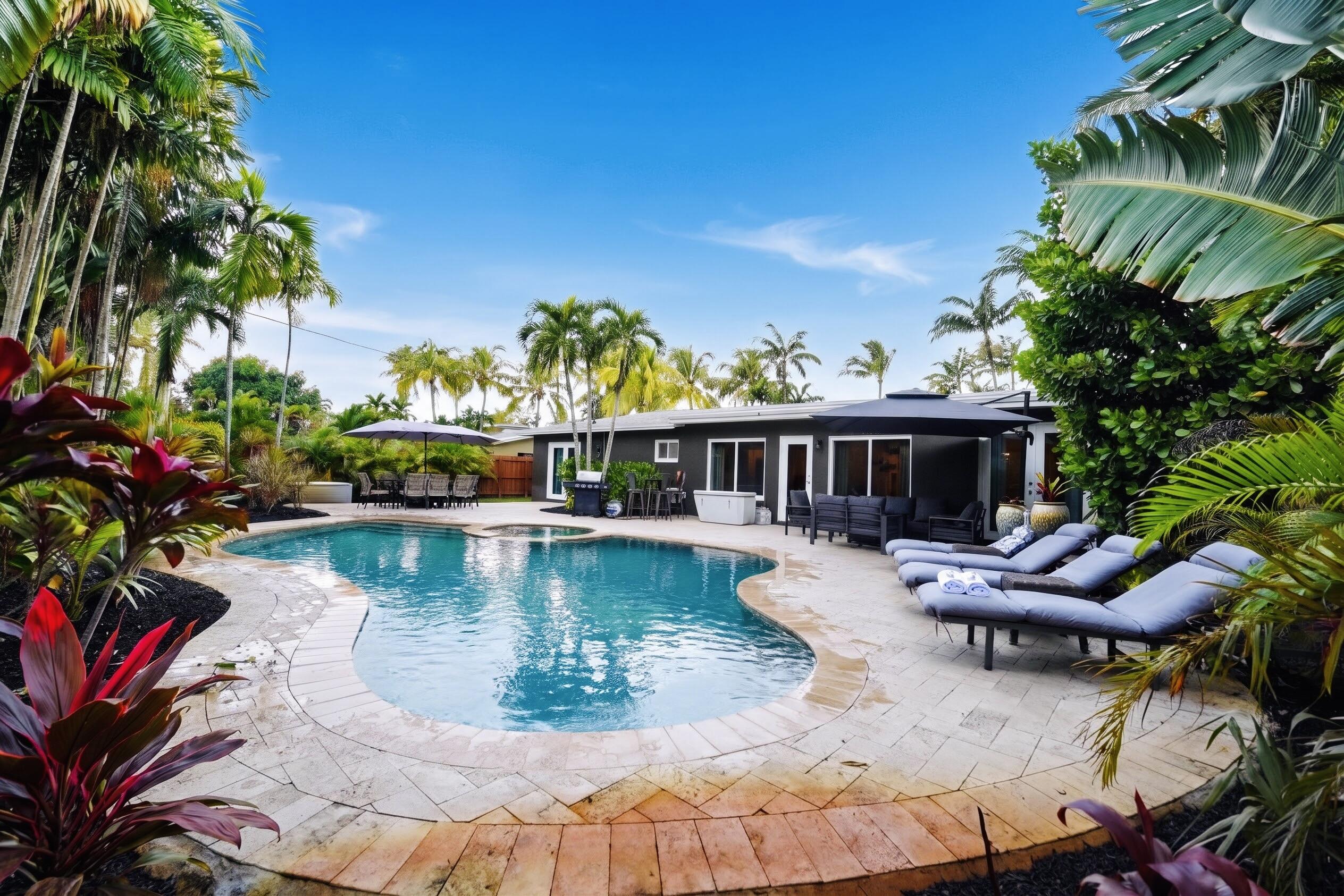 Curved in-ground swimming pool with an integrated spa, surrounded by natural stone hardscaping, mature tropical landscaping, and a dark-toned exterior home facade