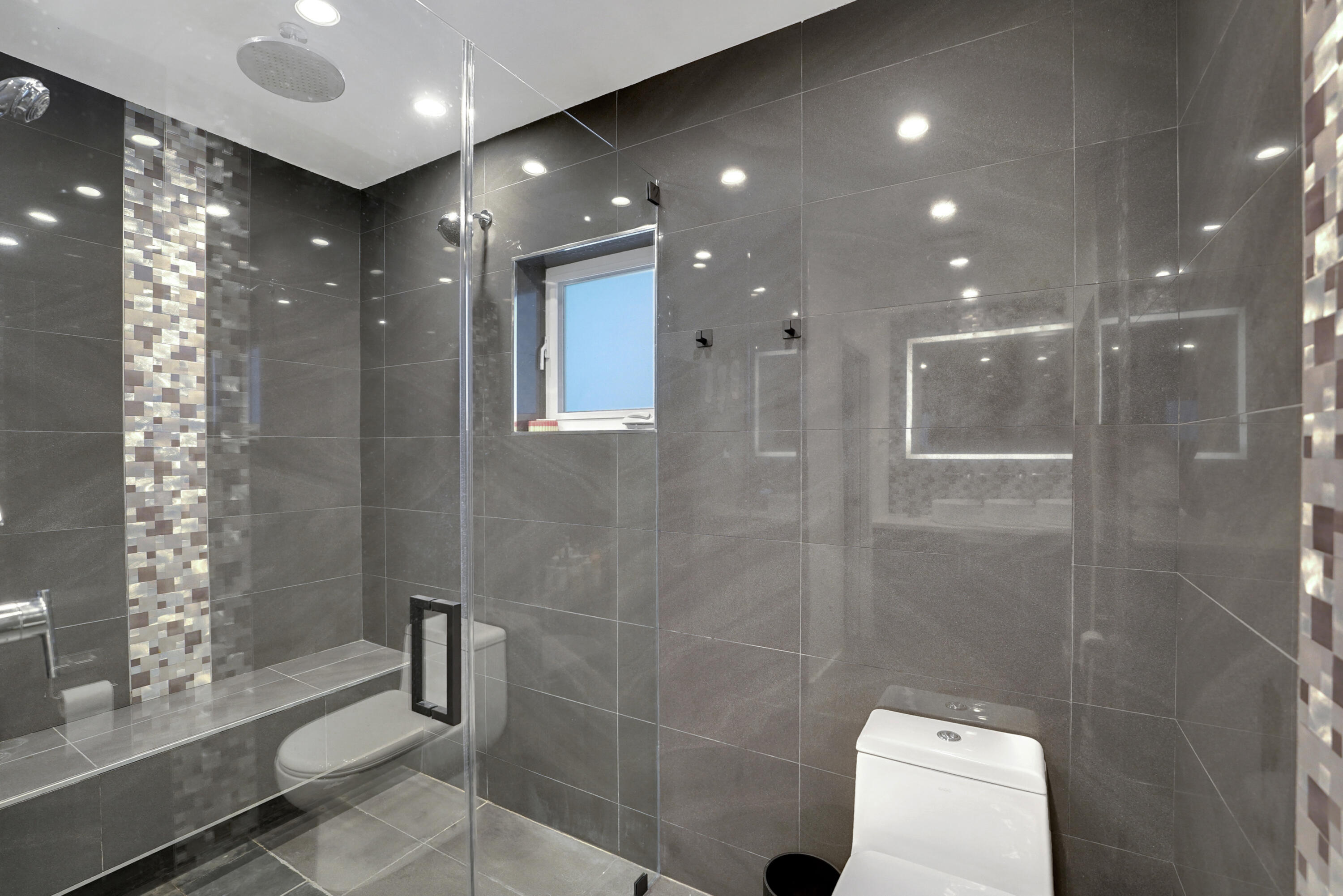 Walk-in shower featuring large-format gray tile, a vertical mosaic accent inlay, a built-in bench, a ceiling-mounted rainfall showerhead, and a frameless glass enclosure