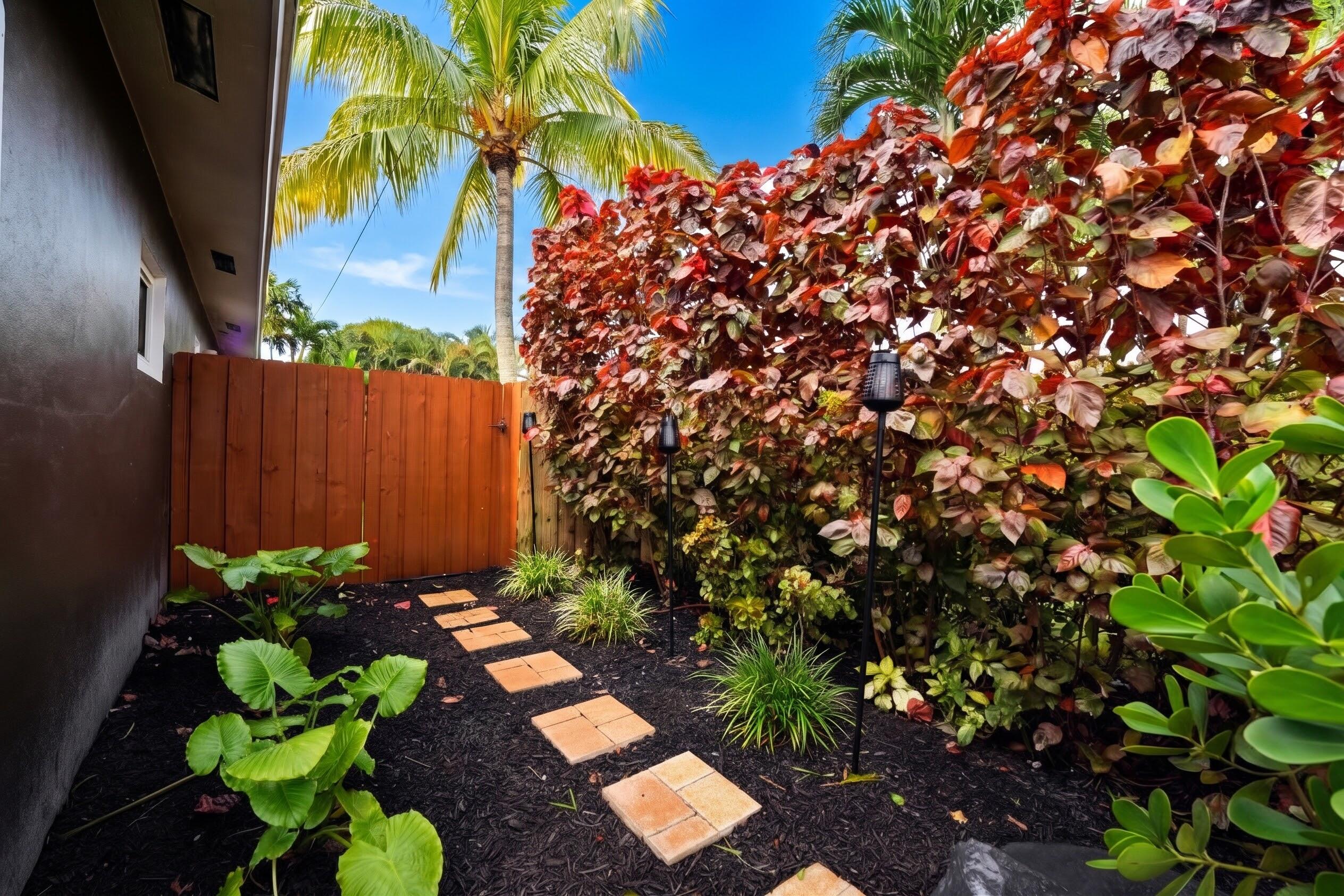 Landscaped side yard featuring a paver stone walkway, dark mulch garden beds, mature tropical plantings, and a wood privacy fence