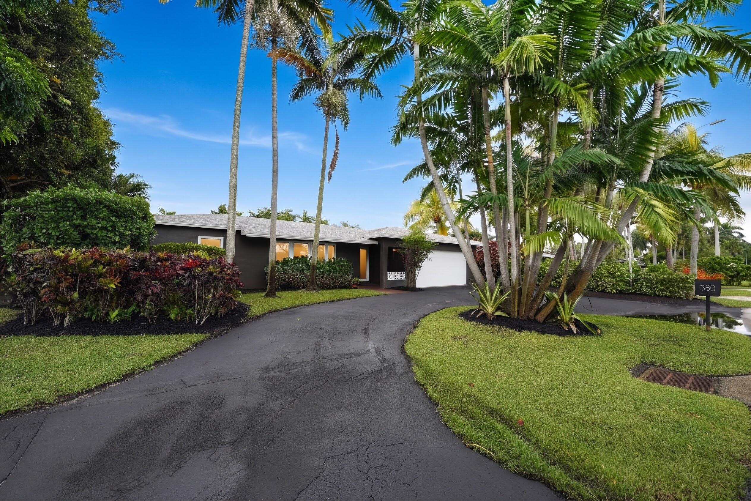 Exterior facade featuring dark-toned siding, attached garage, curved asphalt driveway, and mature palm landscaping with manicured lawn