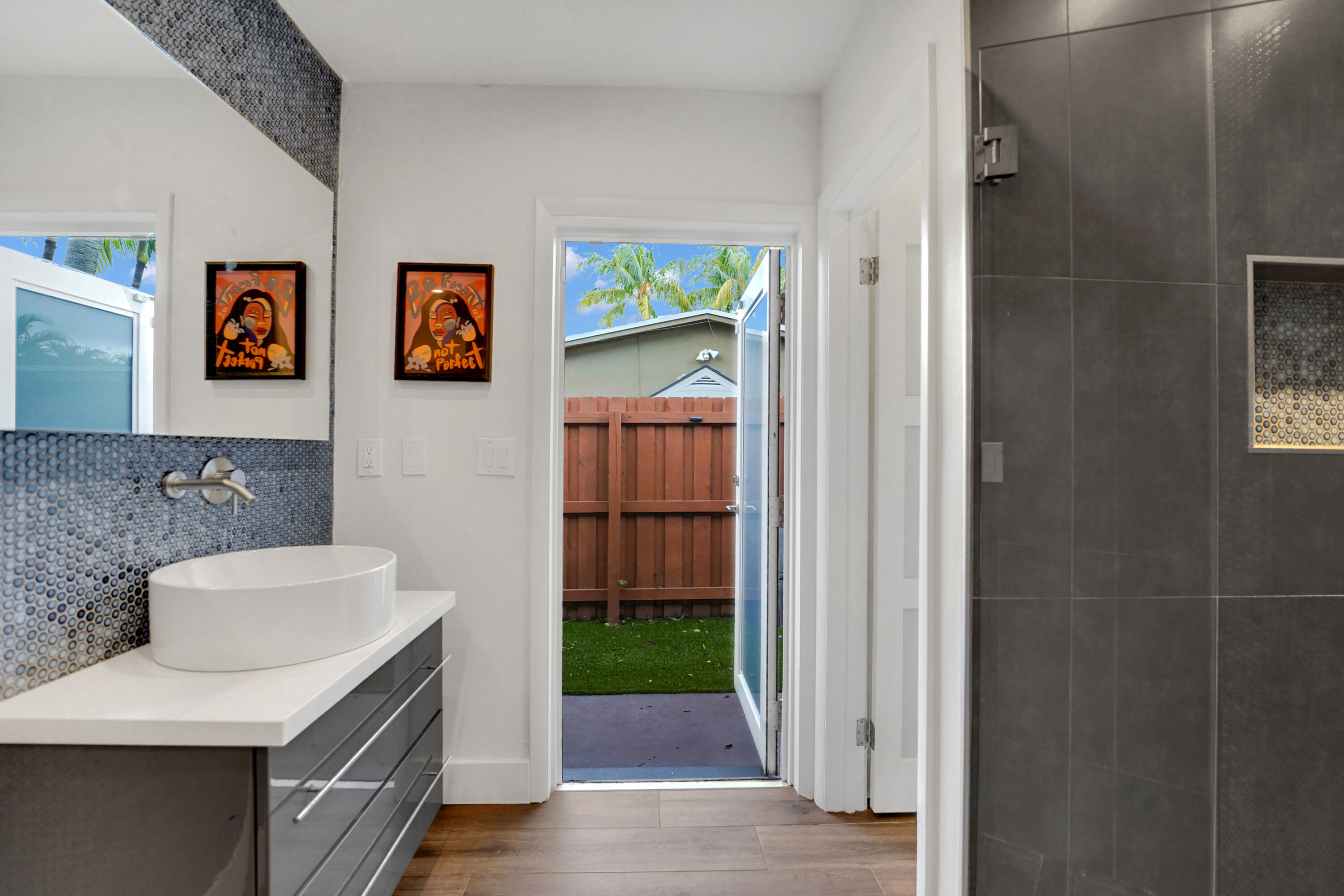 Modern floating vanity with a white vessel sink, quartz countertop, gray high-gloss cabinetry, metallic penny-round mosaic backsplash, and walk-in shower with large-format charcoal tile and a built-in recessed niche