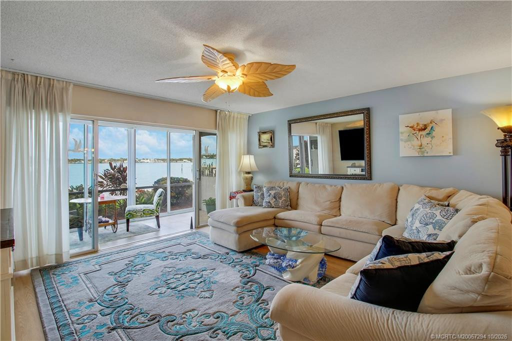 Relax in the comfortable living room with a spacious sectional sofa and tranquil coastal colors. Sliding glass doors fill the space with natural light and open to a waterfront patio overlooking the sparkling blue water, creating a wonderful connection between indoor and outdoor living.